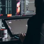 A person sitting at a desk with two monitors, one displaying code and the other displaying a graphical user interface