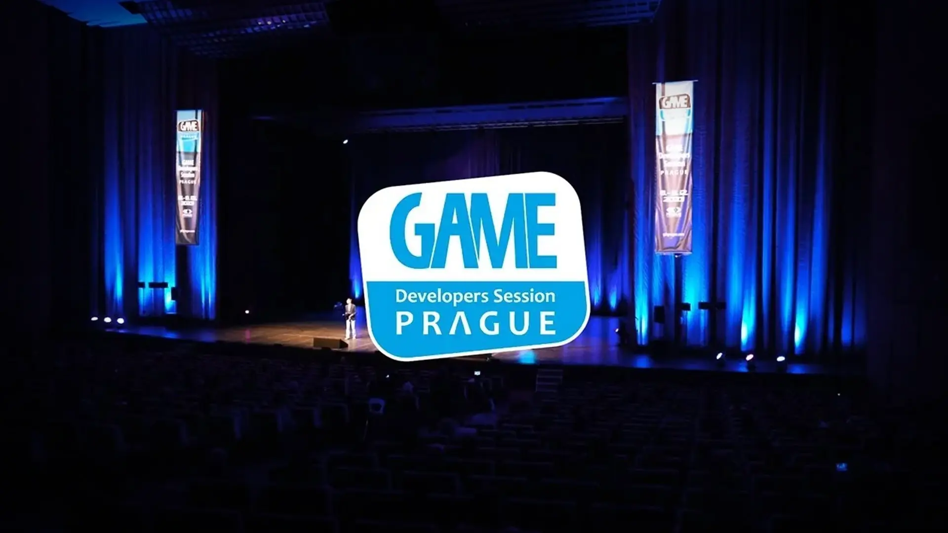 The "GAME Developers Session PRAGUE" logo is prominently displayed over a dimly lit auditorium where a speaker is on stage, with blue lighting on the curtains and banners