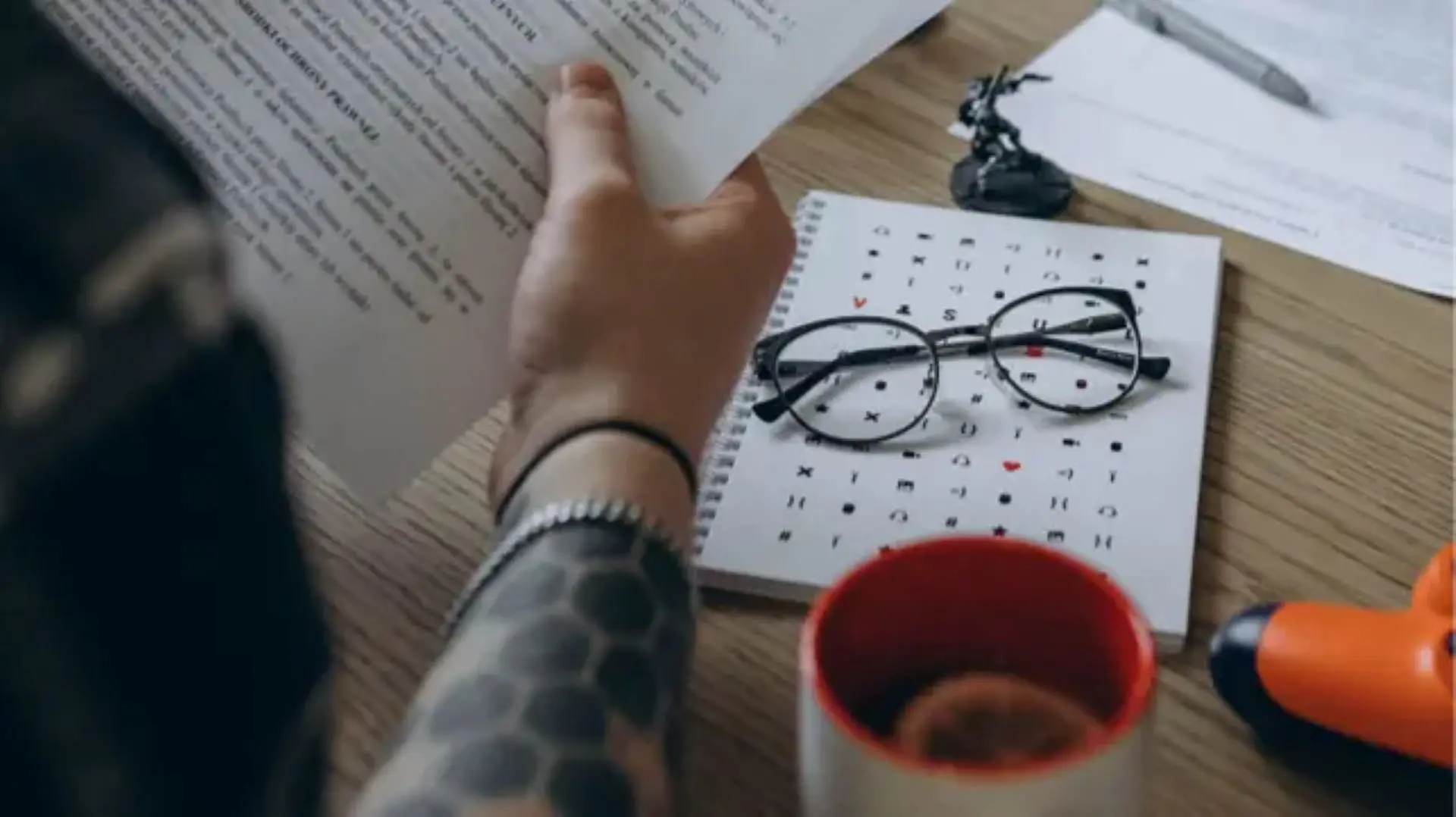 A fragment of a desktop. The hands of a person holding sheets of paper with text are visible. On the table are glasses with a black frame on a notebook with drawings, a pen, a small figurine