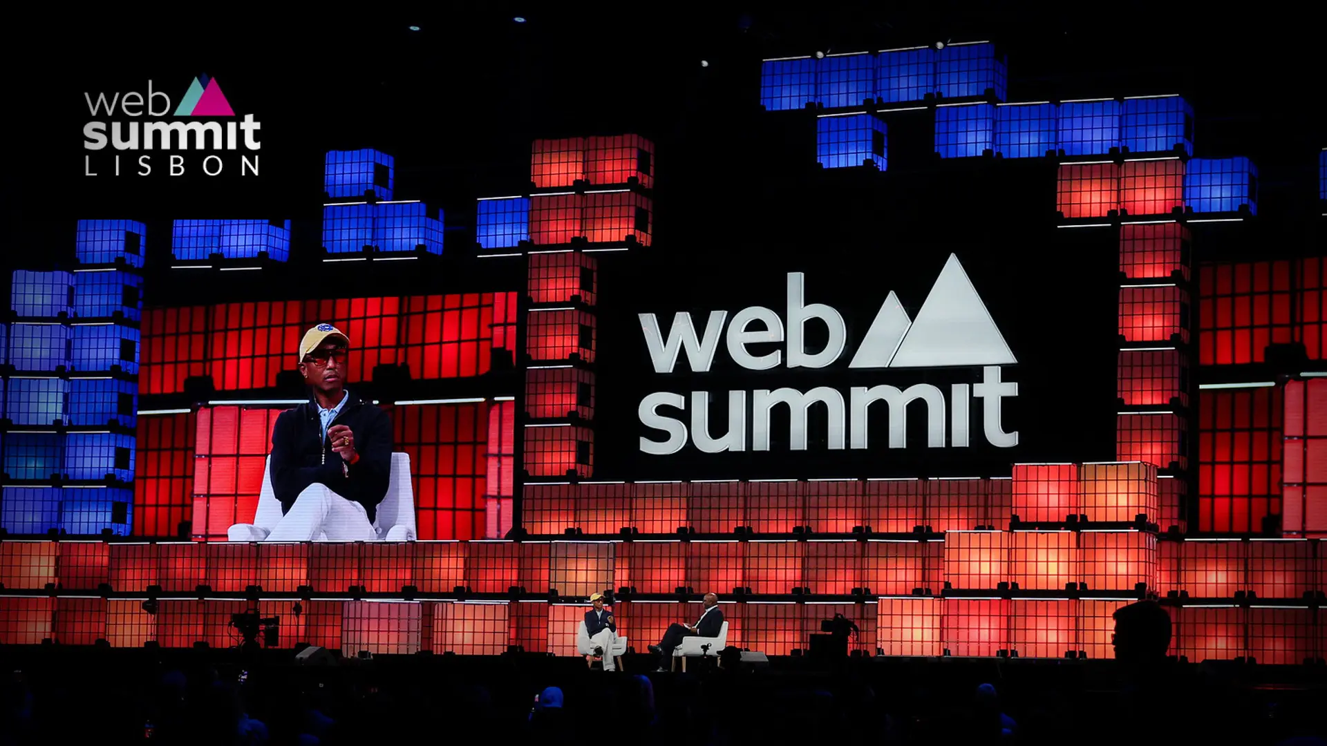 Pharrell Williams is displayed on a large screen above two speakers on stage at the Web Summit Lisbon, which features a backdrop of red and blue illuminated squares and the event's logo.